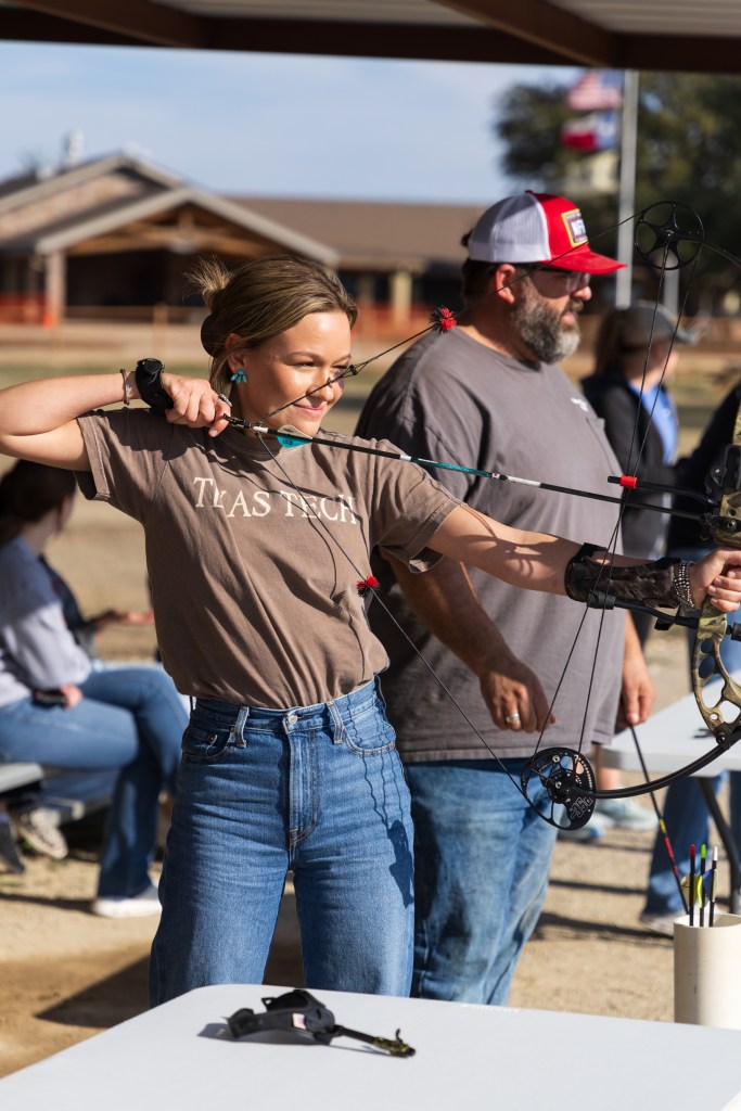 A participant at the MILE retreat confidently draws a bow with a focused expression, preparing to shoot, while others observe in the background.