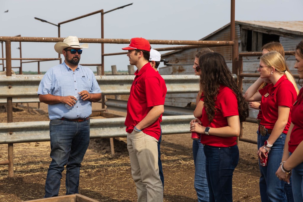 Group of students in red shirts engaged in conversation with a man in a cowboy hat and sunglasses at a ranch in Texas.