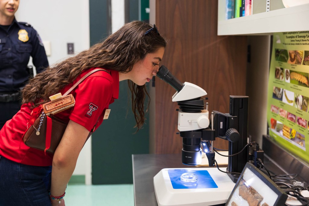 A student in a red shirt examines a sample under a microscope in a laboratory setting, with a police officer in the background.