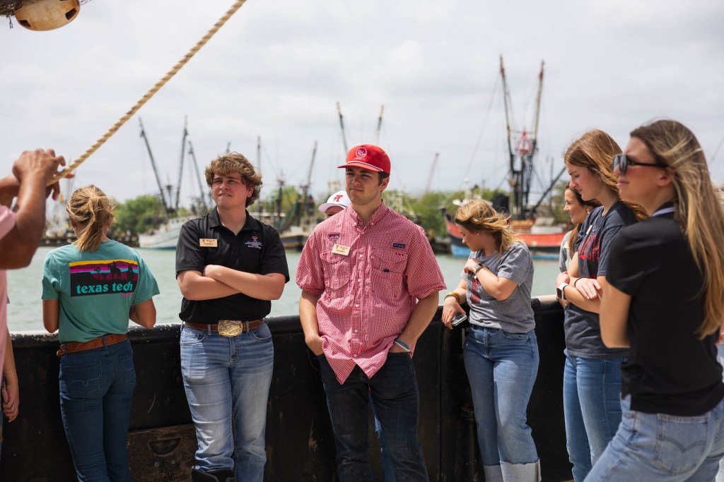 Students from the Matador Institute of Leadership Engagement observing the Rio Grande Valley agriculture industry, featuring various students attentively listening on a dock with fishing boats in the background.
