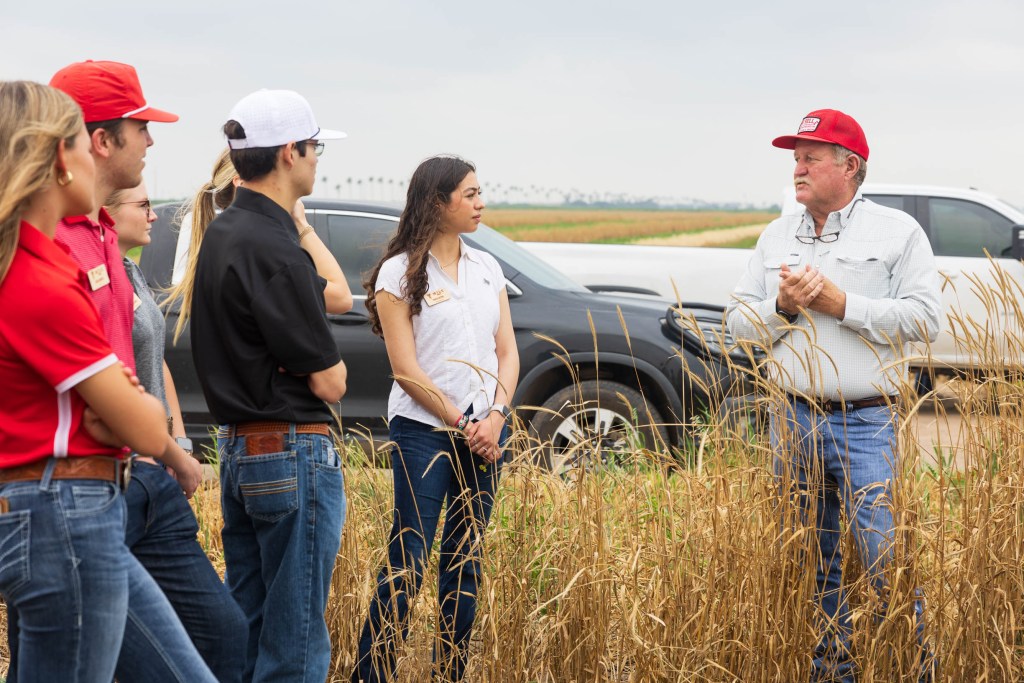 A group of students wearing agricultural attire listens attentively to a speaker standing in a field of tall crops, with vehicles parked in the background.