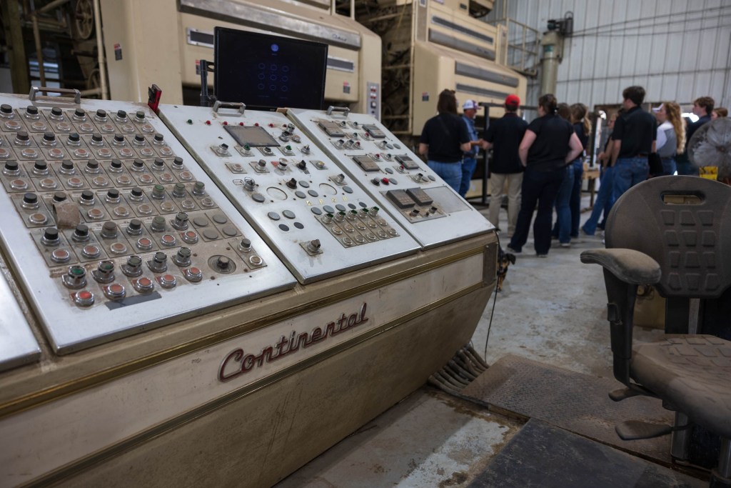 Control panel with numerous buttons and screens in an agricultural facility, with people in the background.