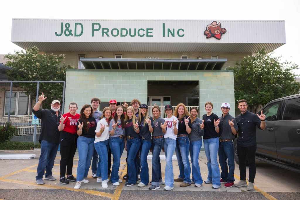 Group photo of students and instructors from the Matador Institute of Leadership Engagement in front of J&D Produce Inc. building, with individuals making a hand gesture for 'hook 'em horns.'