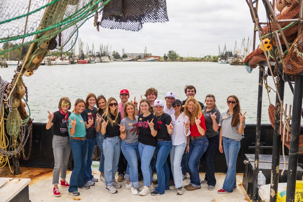 Group photo of MILE program students on a boat in the Rio Grande Valley, posing with smiles and various hand signs against a backdrop of the water and fishing boats.