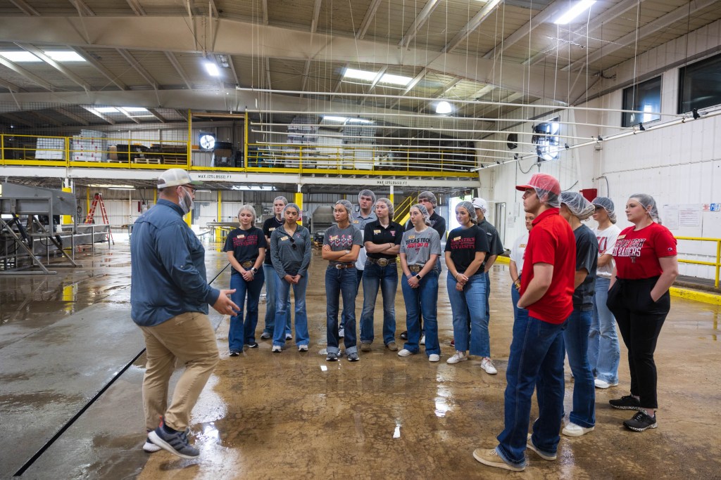 A group of students listening attentively to a speaker during a tour in an agricultural facility, with various industrial equipment visible in the background.