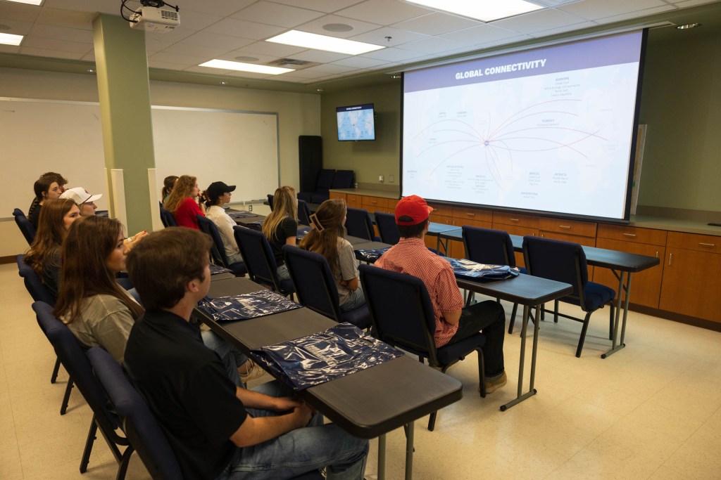 Students attending a presentation about global connectivity in a classroom setting, with a projection screen displaying a map and lines indicating connections to various regions.