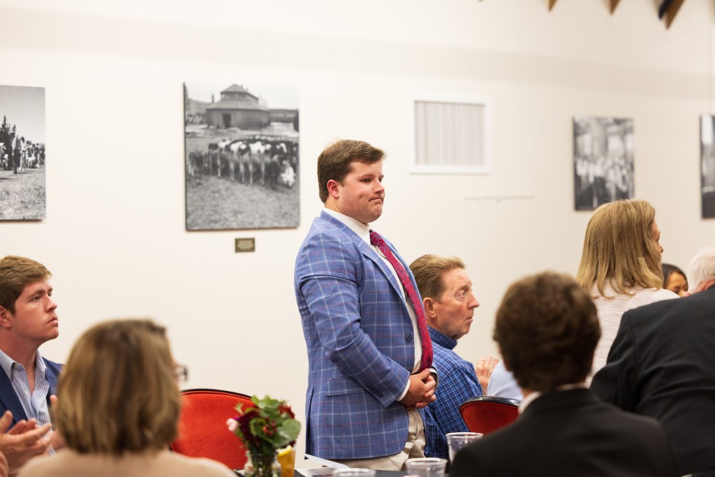 A student wearing a blue plaid blazer stands during a graduation ceremony at Texas Tech University's Dairy Barn, with attendees seated around tables in the foreground.