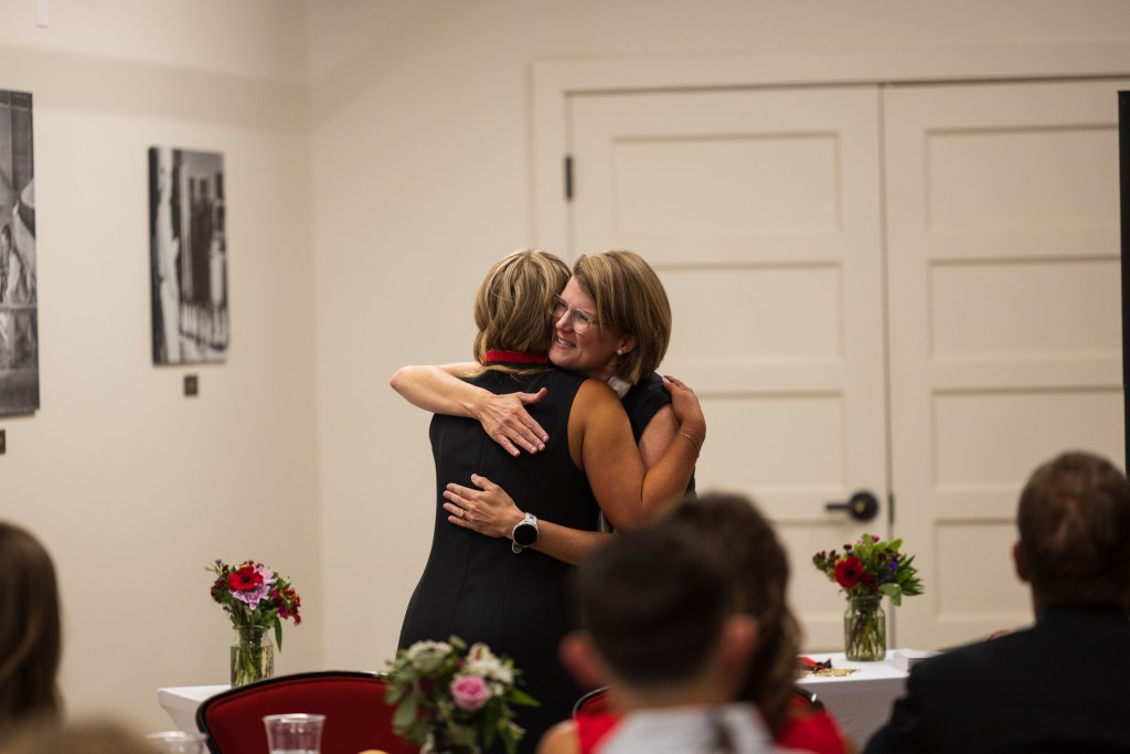 Two women embrace in celebration during a graduation ceremony, with flowers on the table in the foreground and historical photographs on the wall behind them.