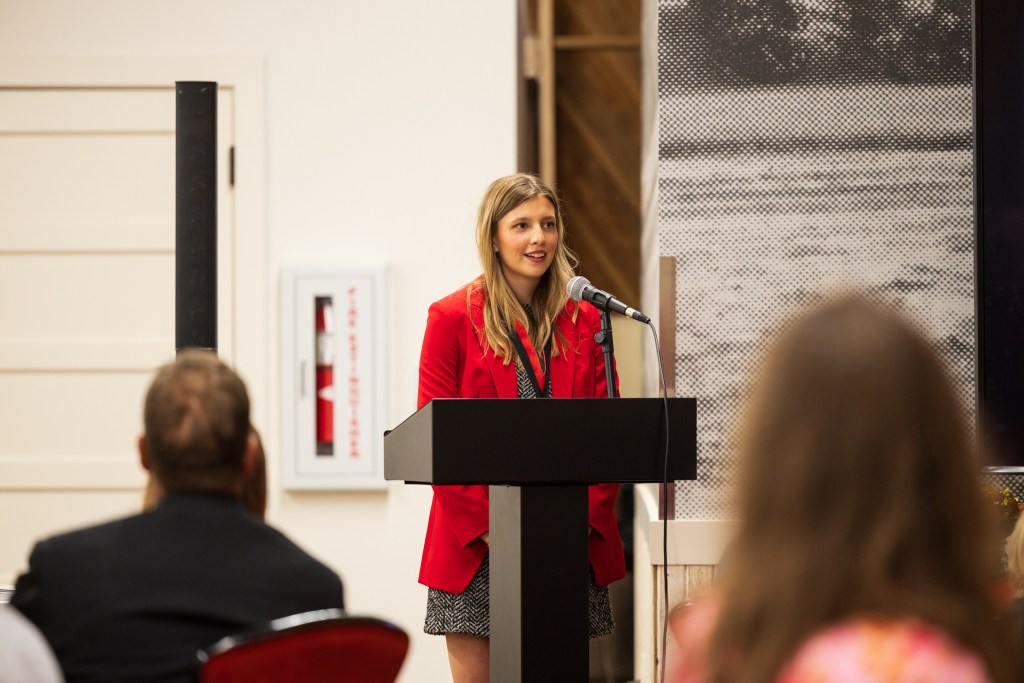 A young woman in a red blazer speaks at a podium during a graduation ceremony at Texas Tech University, with audience members listening attentively in the foreground.