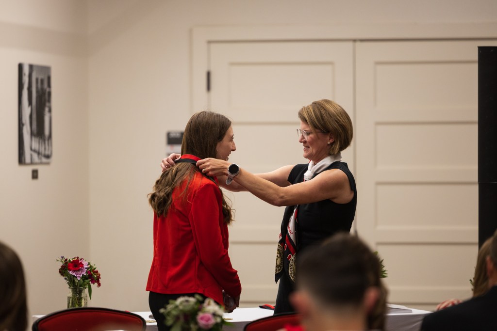 A woman in a red jacket receives a medal during a graduation ceremony, while another woman in a black dress assists her. The setting features tables with floral arrangements and a background of historical photographs.