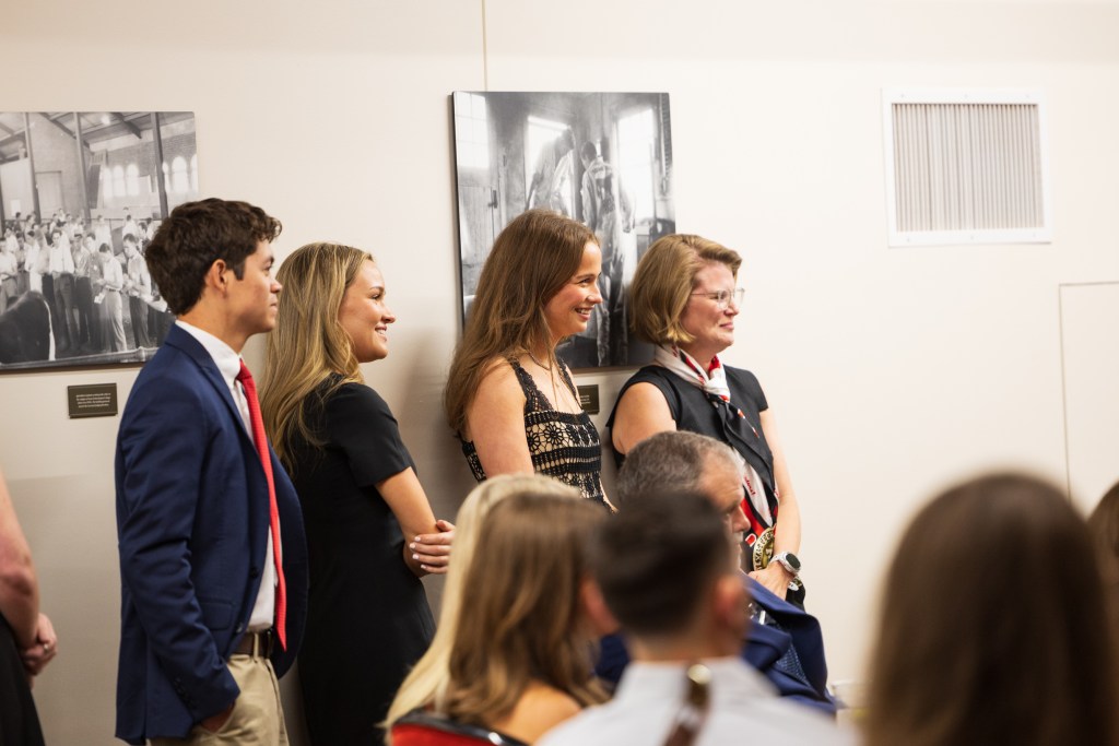 Group of four individuals standing together during a graduation ceremony, smiling and engaged, with historical black and white photographs displayed on the wall behind them.