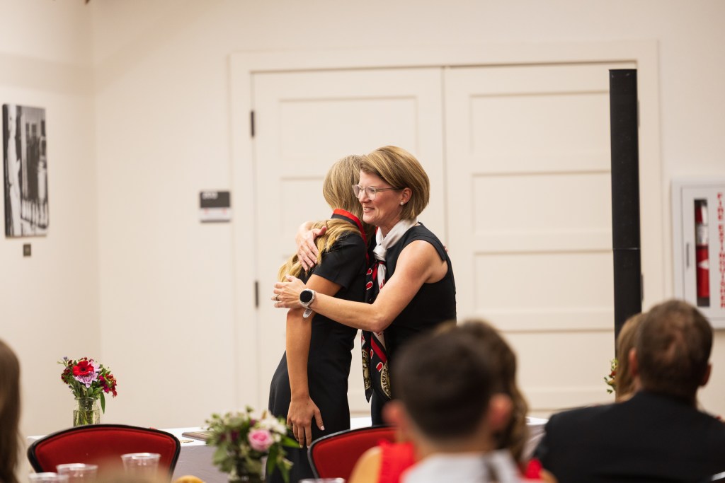 A graduate and a program director embrace during the MILE Program graduation ceremony at Texas Tech University, surrounded by decorated tables and floral arrangements.
