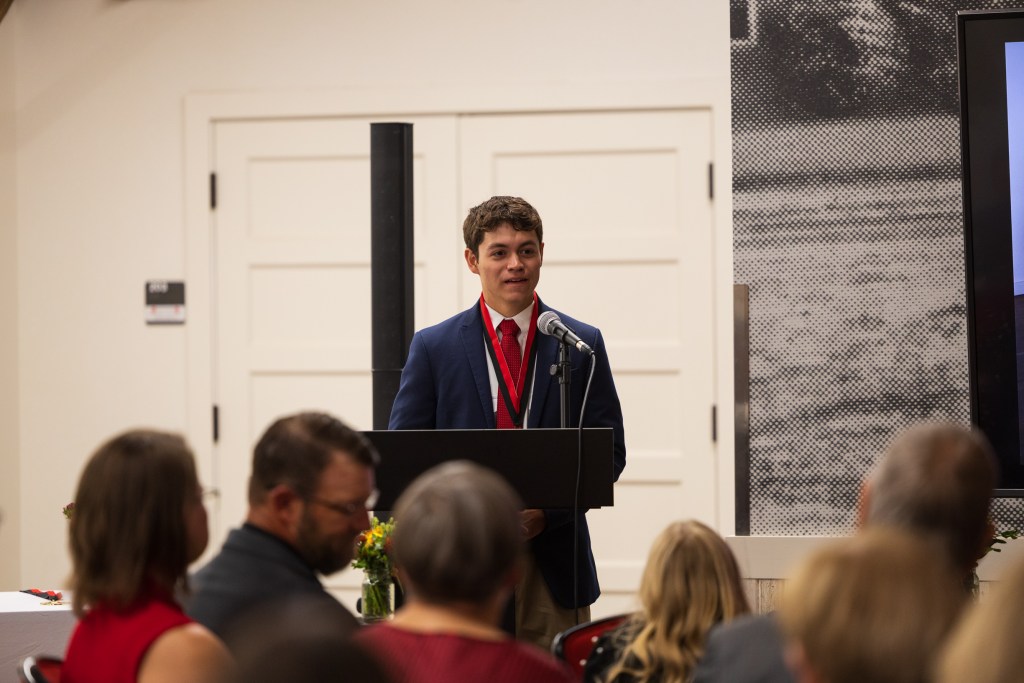 A student giving a speech during the graduation ceremony for the MILE Program at Texas Tech University, standing at a podium with a microphone and addressing the audience.
