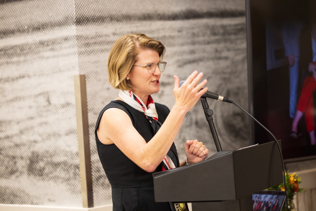 A woman speaking at a podium during a graduation ceremony, wearing glasses and a black dress with a decorative scarf.