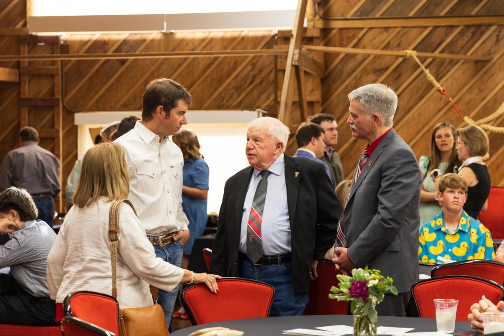 Guests engage in conversation during the MILE Program graduation ceremony at Texas Tech University, featuring a rustic wooden background and tables with floral centerpieces.