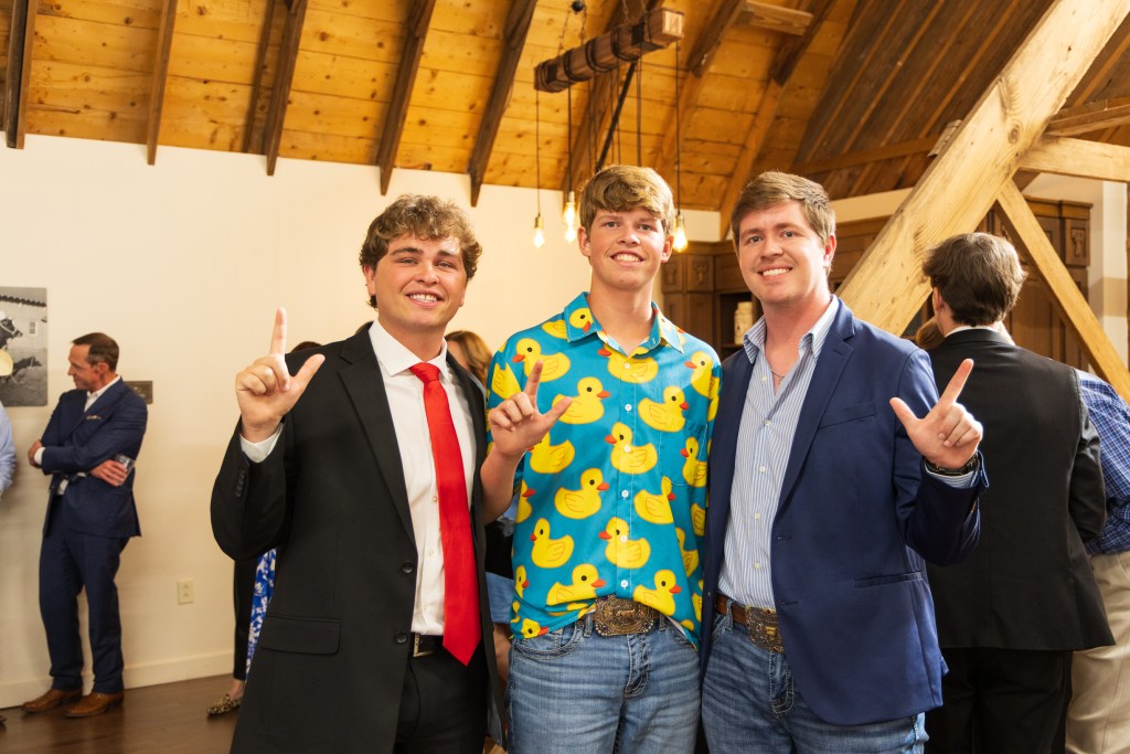 Three male graduates celebrating at the MILE Program graduation ceremony, with one wearing a bright duck-patterned shirt. They are posing with hand signs in a rustic venue.
