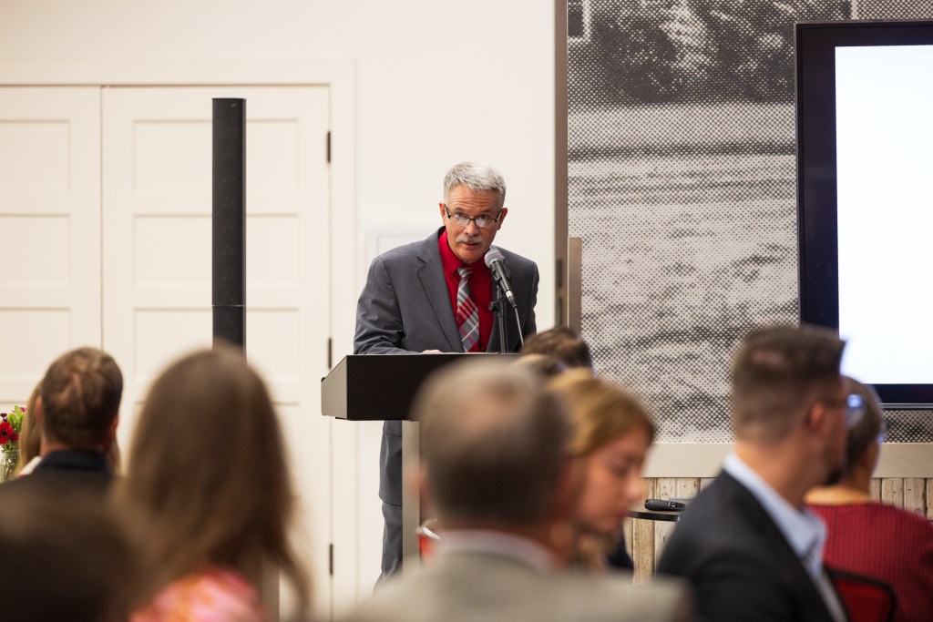 A speaker presenting during a graduation ceremony at the Texas Tech University Dairy Barn, with attendees seated in the foreground.