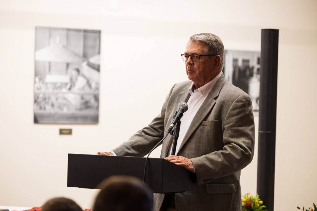 A speaker stands behind a podium during a graduation ceremony, addressing the audience with a serious expression. In the background, historic photographs are displayed on the wall.